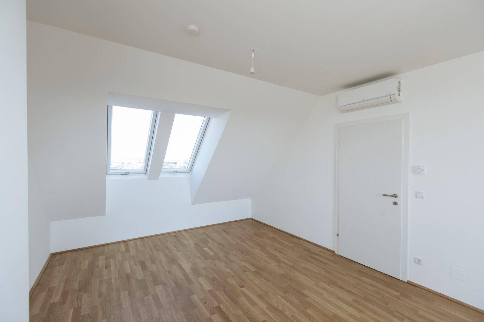 Heller Raum im Dachgeschoss mit Holzboden, zwei Dachfenstern und einer Klimaanlage für Komfort.