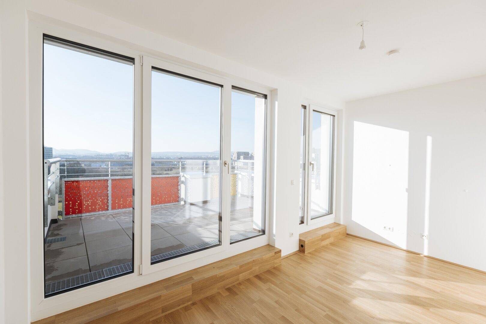 Lichtdurchfluteter Raum mit Holzboden, großen Glasfronten und direktem Zugang zu einer sonnigen Terrasse.