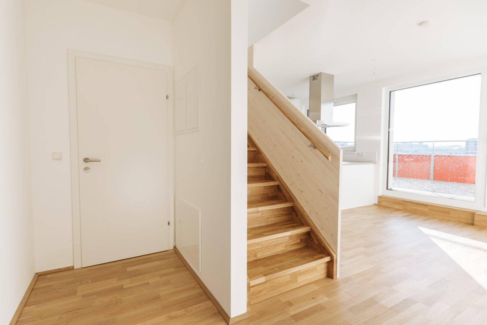 Heller Eingangsbereich mit Holzboden und Holztreppe, offener Blick in die moderne Küche.