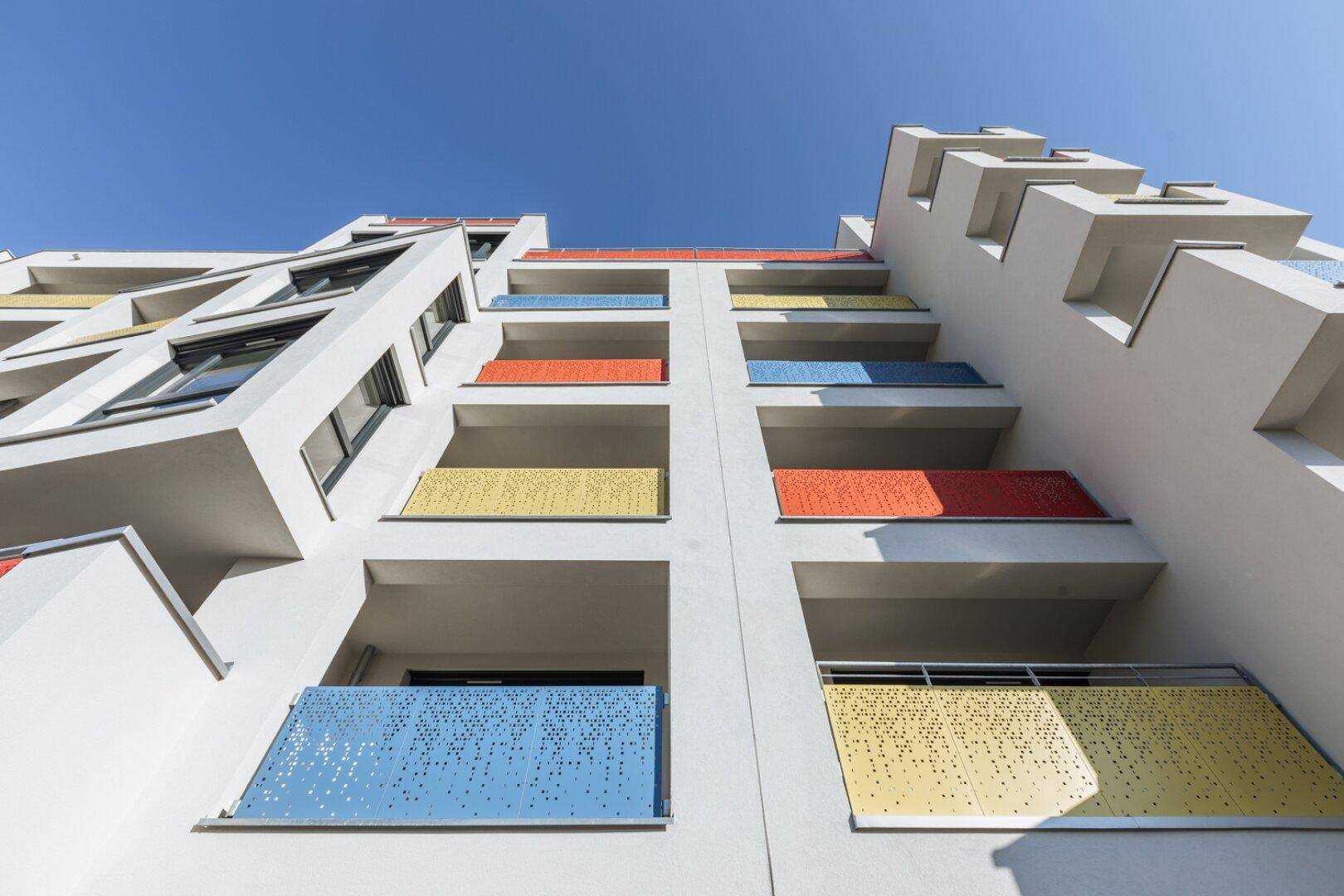 Moderne Gebäudefassade mit farbenfrohen Balkonen in Blau, Orange und Gelb unter blauem Himmel.