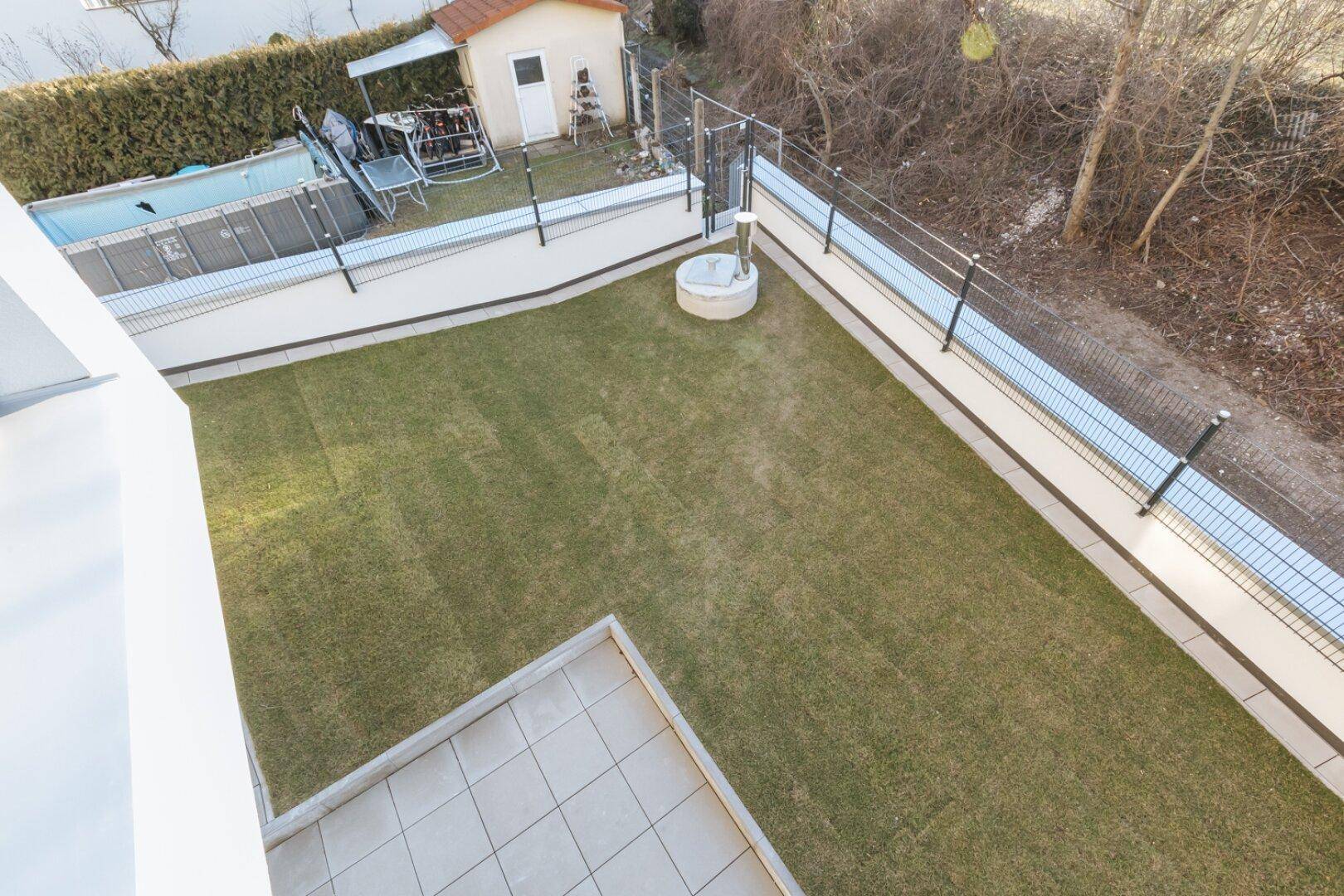 Gepflegter Gartenbereich mit Rasenfläche, kleiner Terrasse und einem Pool im Hintergrund.