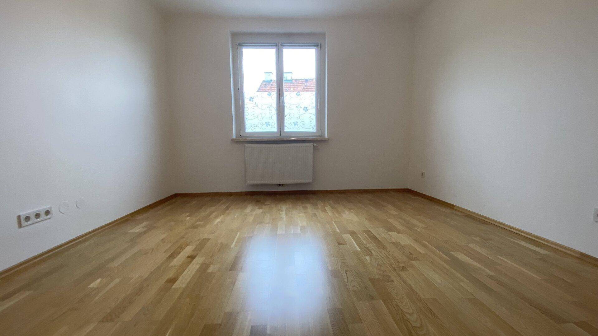 Helles Zimmer mit Holzparkettboden, einem Fenster und Heizkörper, ideal als Schlaf- oder Arbeitszimmer.