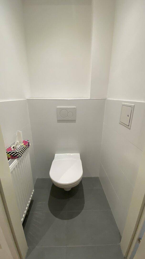Modernes Gäste-WC mit wandhängender Toilette, grauen Fliesen und weiß gefliesten Wänden.