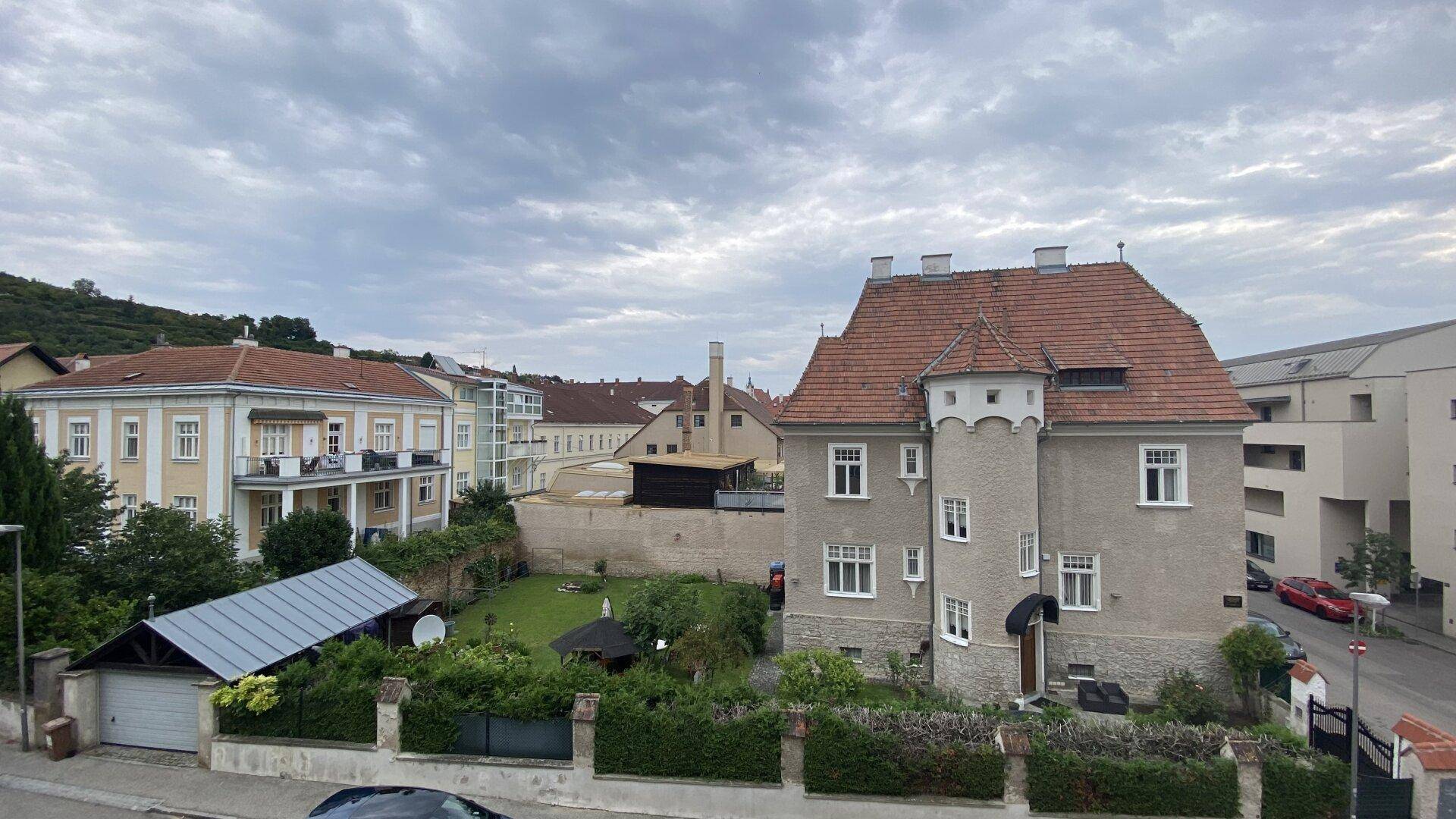 Historisches Gebäude mit Garten und umliegenden Häusern unter bewölktem Himmel.