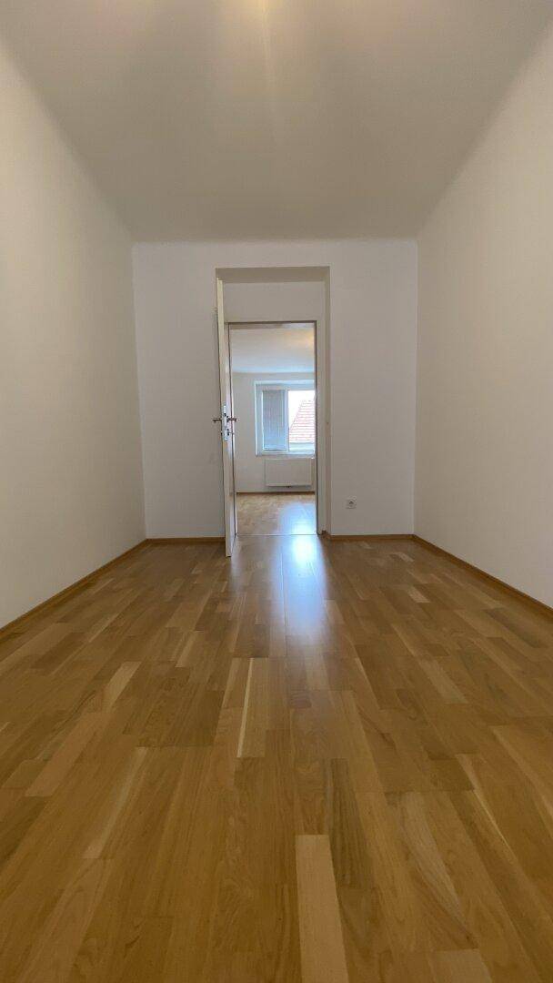 Leeres Zimmer mit Holzparkettboden und weiß gestrichenen Wänden, mit Blick in einen weiteren Raum.
