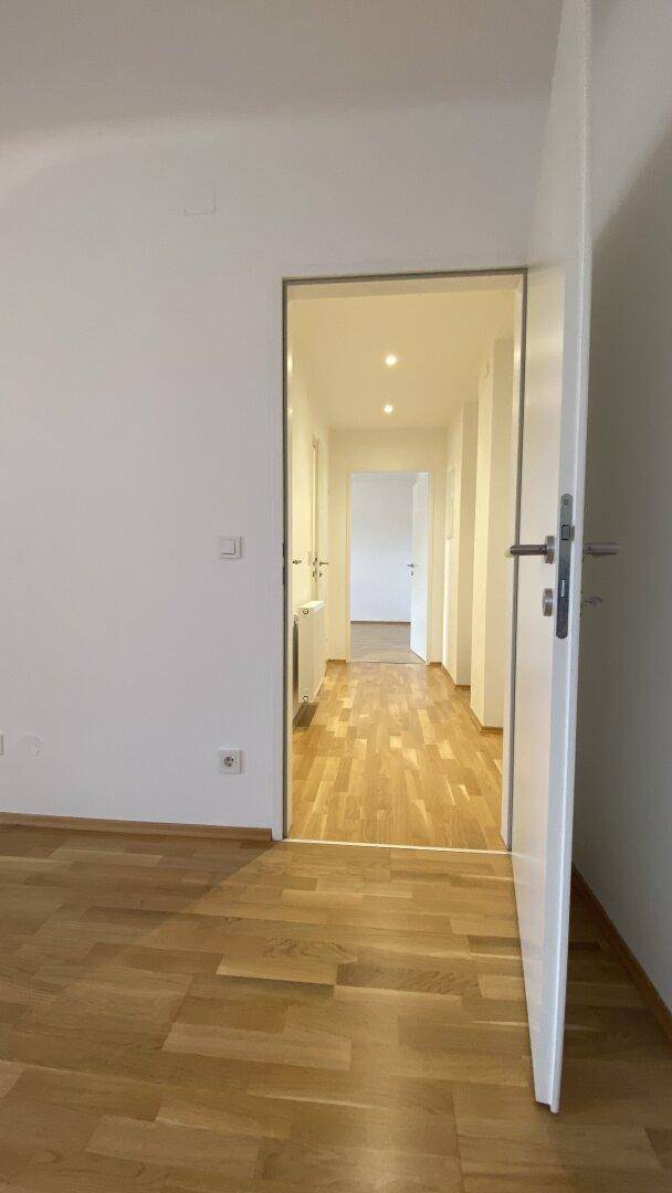 Blick aus einem Zimmer in den hellen Flur mit durchgehendem Holzparkettboden und weißen Wänden.