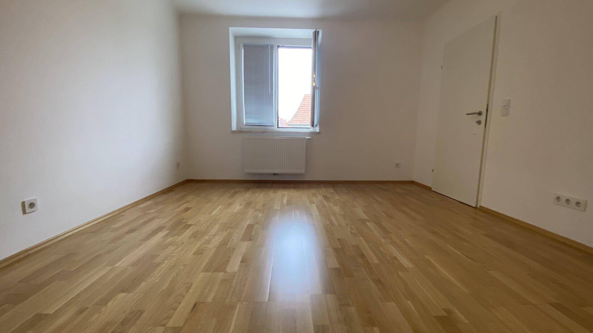 Geräumiges, helles Zimmer mit Holzparkettboden und einem großen Fenster, bereit zur Einrichtung.