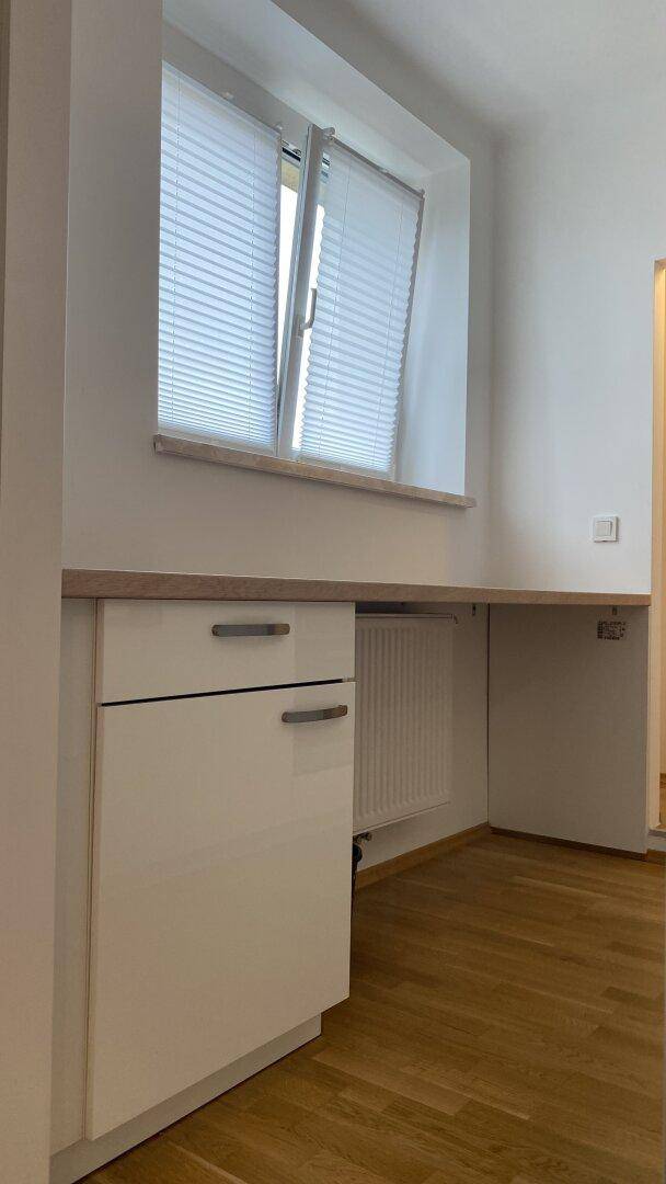 Küchenbereich mit Fenster, weißen Unterschränken und einer Arbeitsplatte, die als Schreibtisch dient.