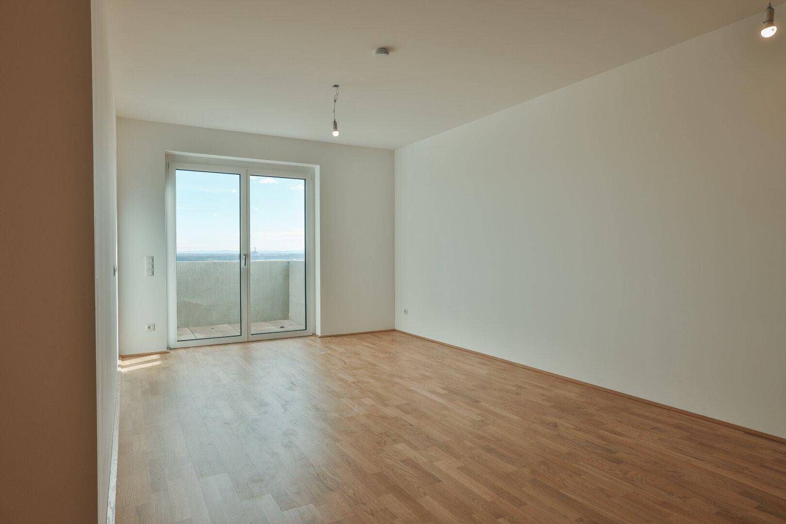 Geräumiges Wohnzimmer mit hellem Parkettboden und Zugang zu einem Balkon mit Weitblick.