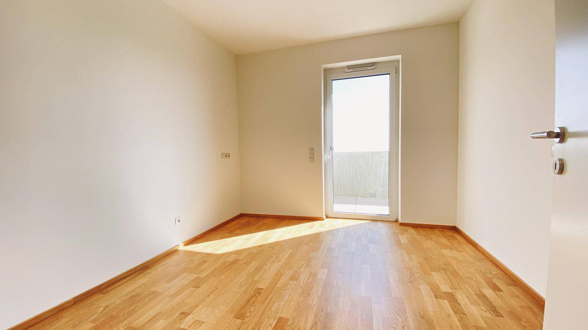 Unmöblierter Raum mit hellem Parkettboden und einer Tür, die auf einen Balkon führt, gut belichtet.