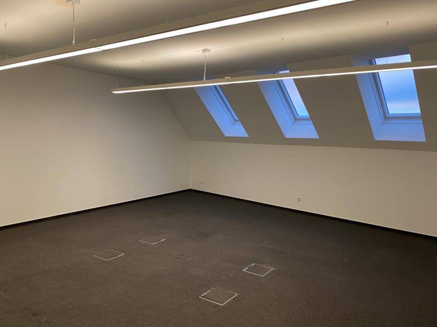 Geräumiger Büroraum mit dunklem Teppichboden, Dachschrägen und mehreren Dachfenstern für viel Tageslicht.