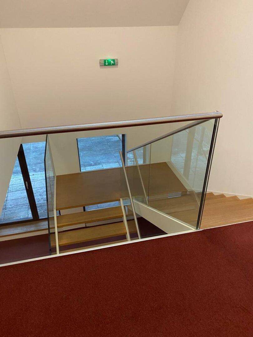 Blick von oben auf einen modernen Treppenaufgang mit Glasgeländer und rotem Teppichboden in einem hellen Flur.