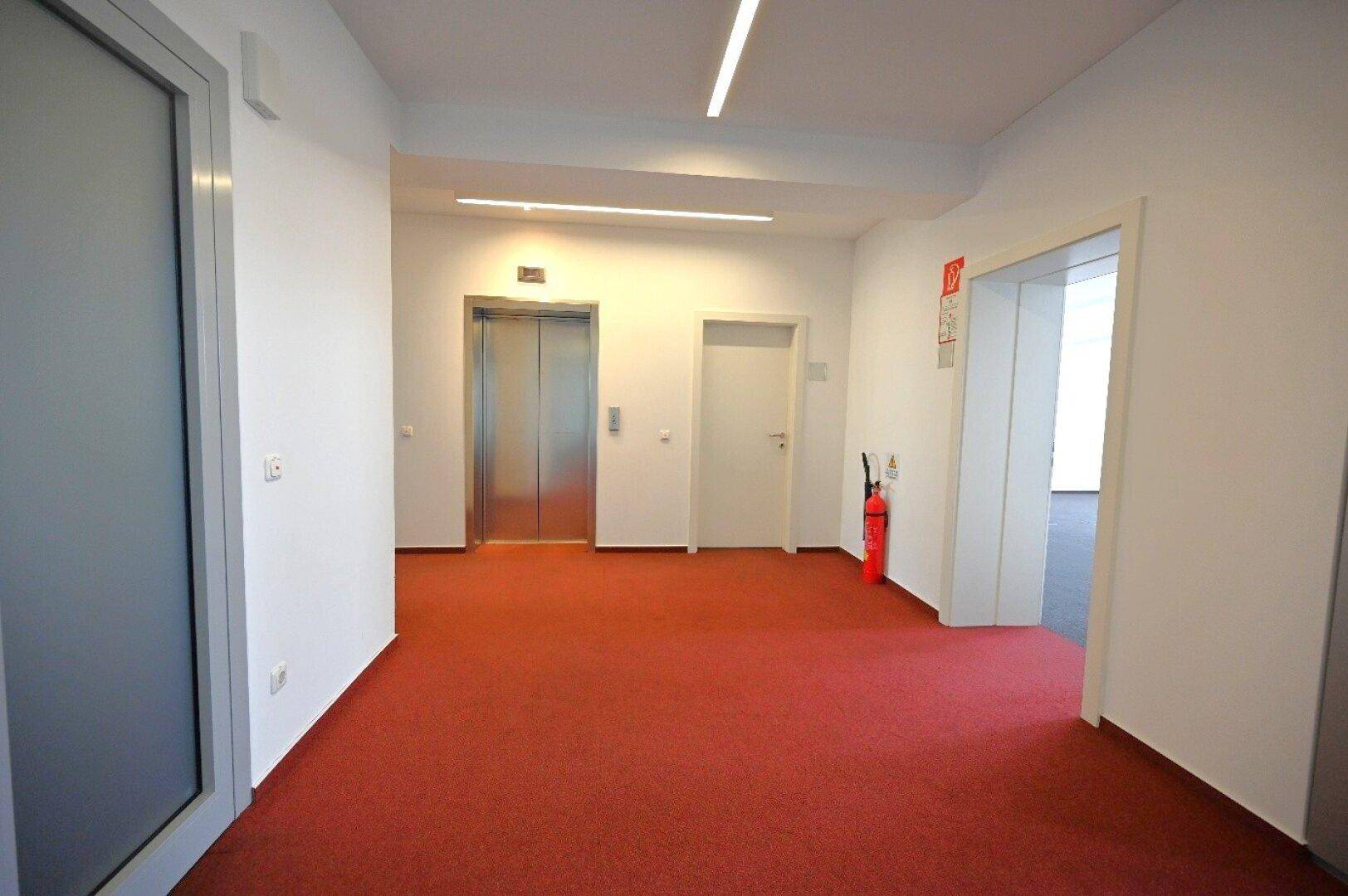 Heller Flur mit rotem Teppichboden und zwei modernen Aufzügen, der Zugang zu den Büroräumen bietet.