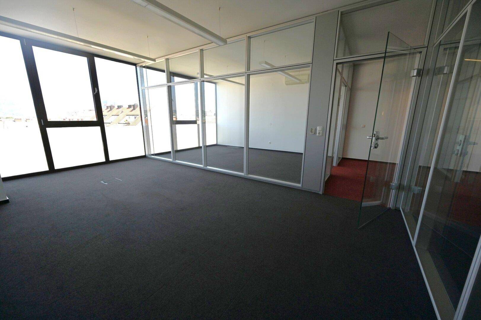 Großzügiger Bürobereich mit dunklem Teppichboden, raumhohen Fenstern und modernen Glaswänden für separate Büros.
