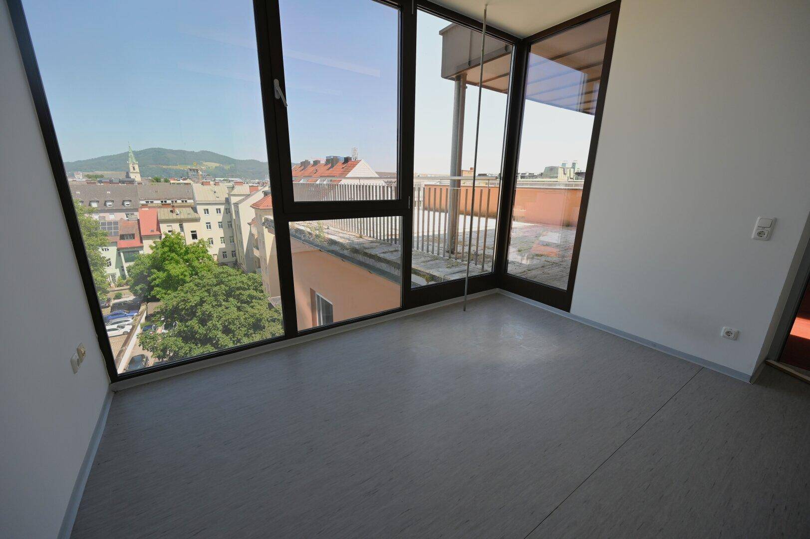 Heller Büroraum mit grauem Bodenbelag und großen Eckfenstern, die einen weiten Blick über die Stadt bieten.