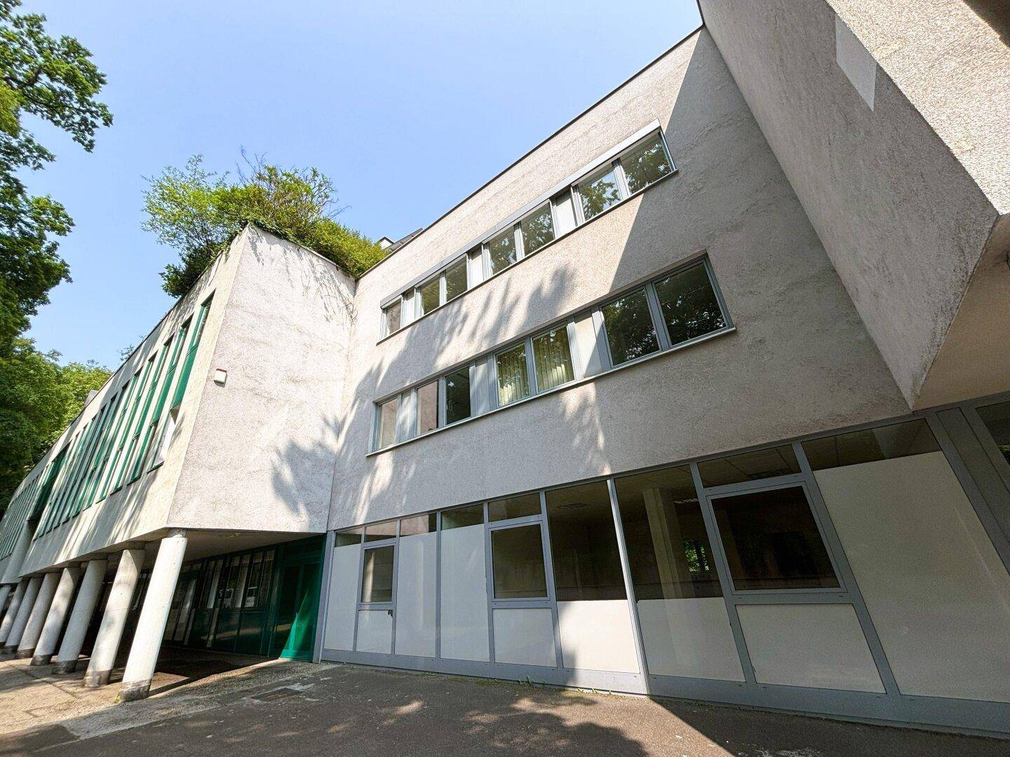 Moderne Architektur des Gebäudes mit großen Fenstern, Säulen und begrüntem Dachbereich.
