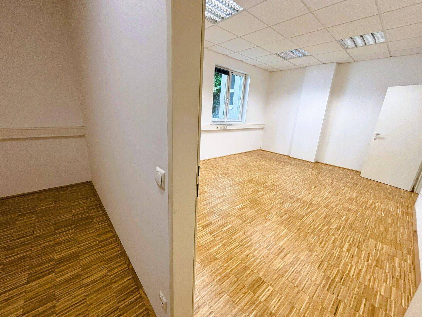 Leerer Büroraum mit Parkettboden, weißen Wänden und einem Fenster, das Tageslicht hereinlässt.