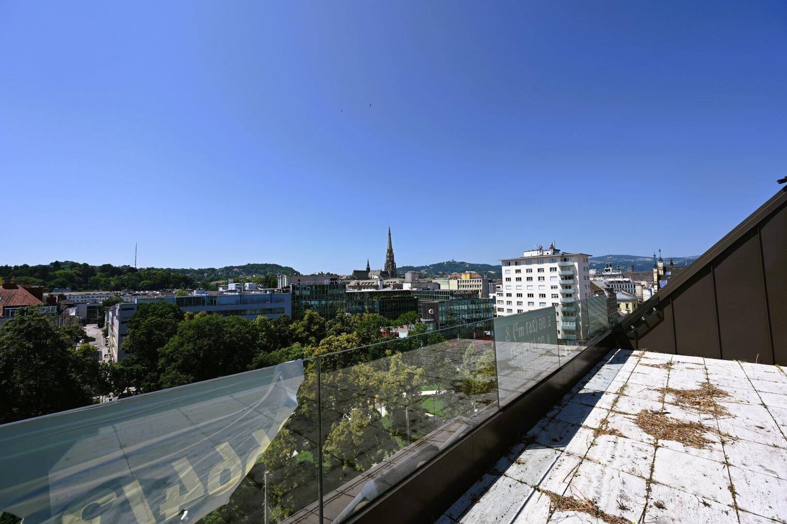 Panoramablick über die Stadt von einem Dach, mit Gebäuden, Bäumen und fernen Bergen unter blauem Himmel.