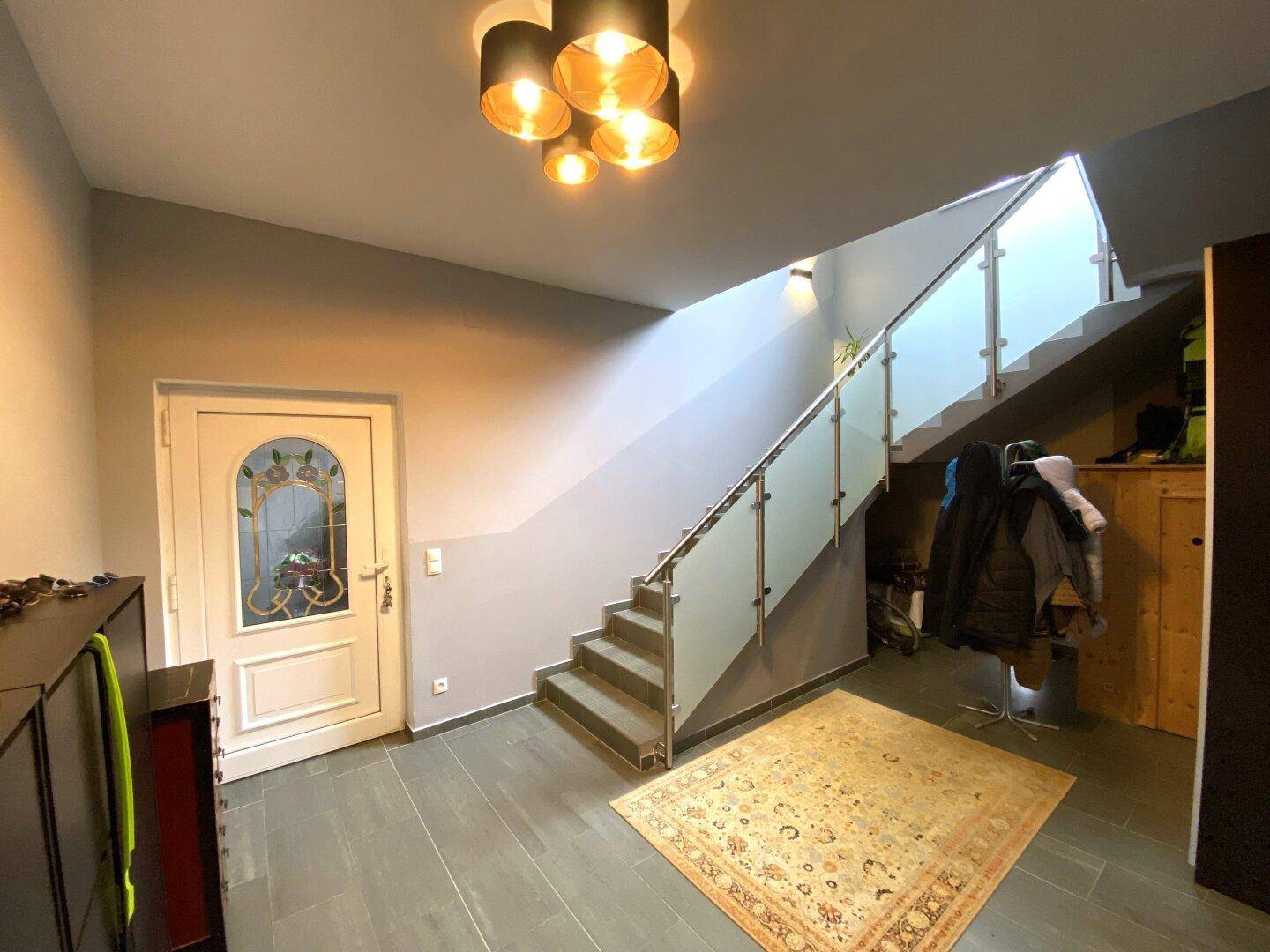 Heller Eingangsbereich mit moderner Beleuchtung, Fliesenboden und Treppe mit Glasgeländer.