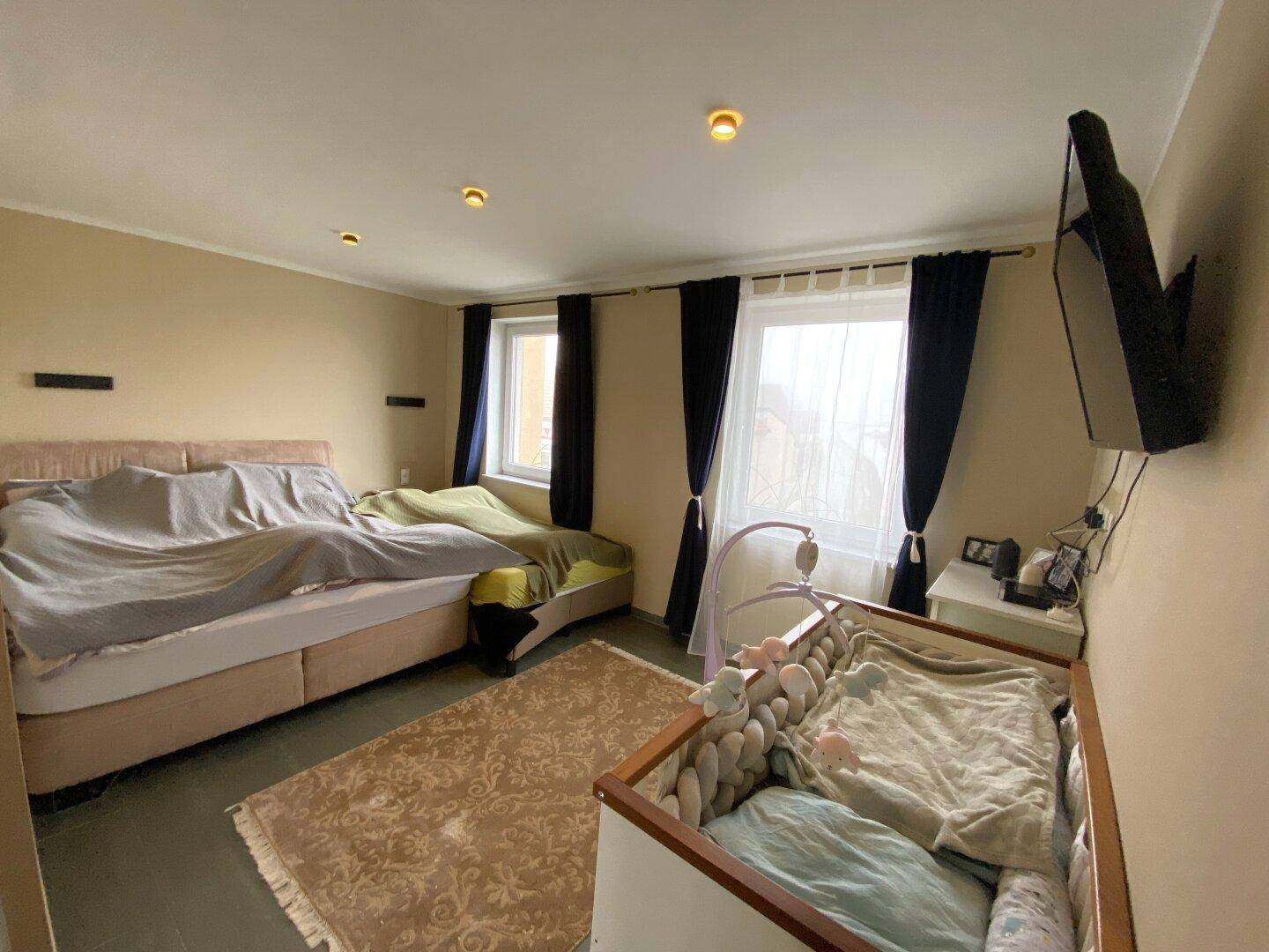 Schlafzimmer mit Doppelbett, Babybett und zwei Fenstern für natürliche Belichtung.