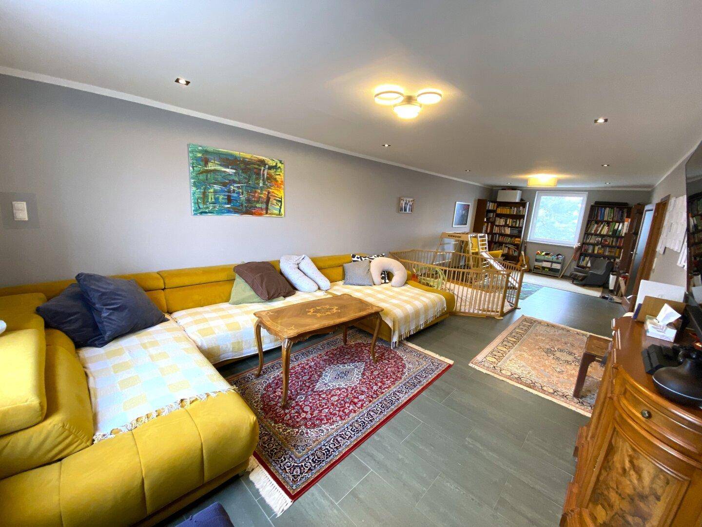 Geräumiges Wohnzimmer mit gelbem Ecksofa, Teppich und Blick auf den Bücherbereich.