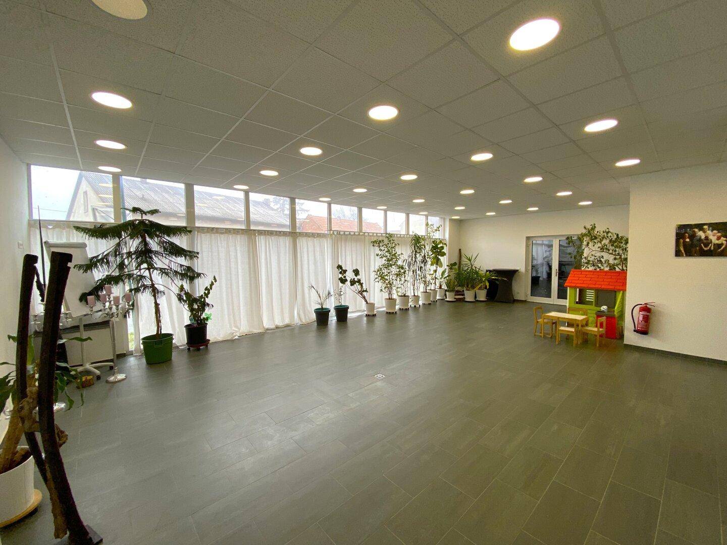 Großer Mehrzweckraum mit vielen Fenstern, Pflanzen und Spielbereich für Kinder.