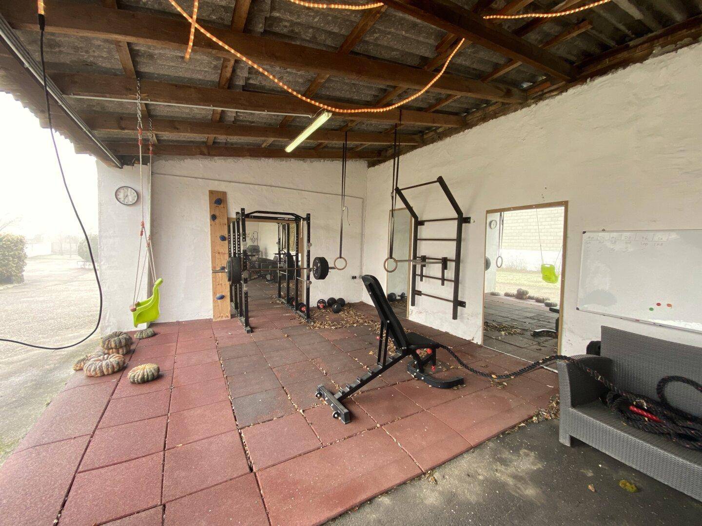 Überdachter Fitnessbereich im Freien mit Trainingsgeräten und Gummiboden.