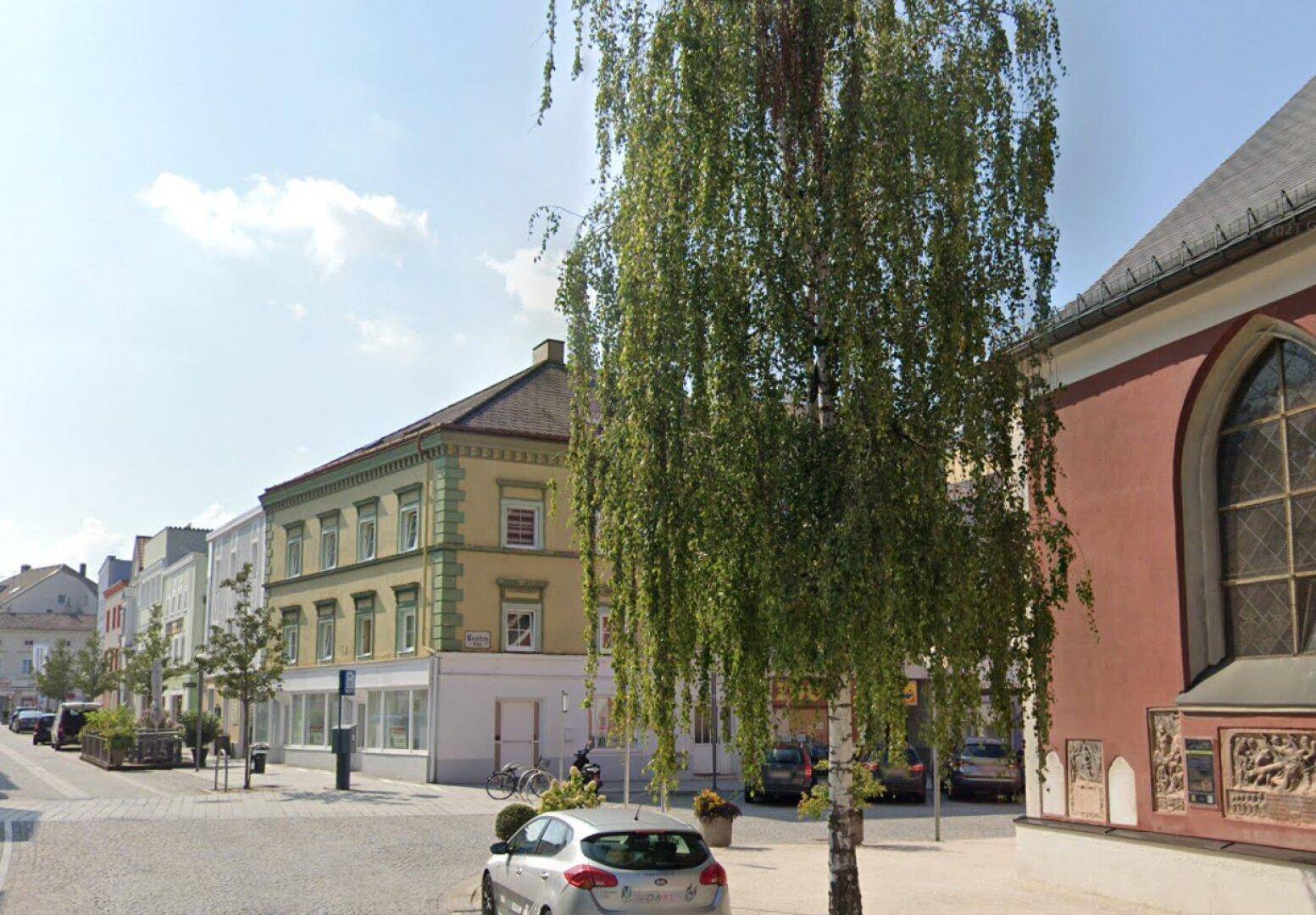 Blick auf eine historische Straße mit Gebäuden, einem großen Baum und einem Teil der Kirche.