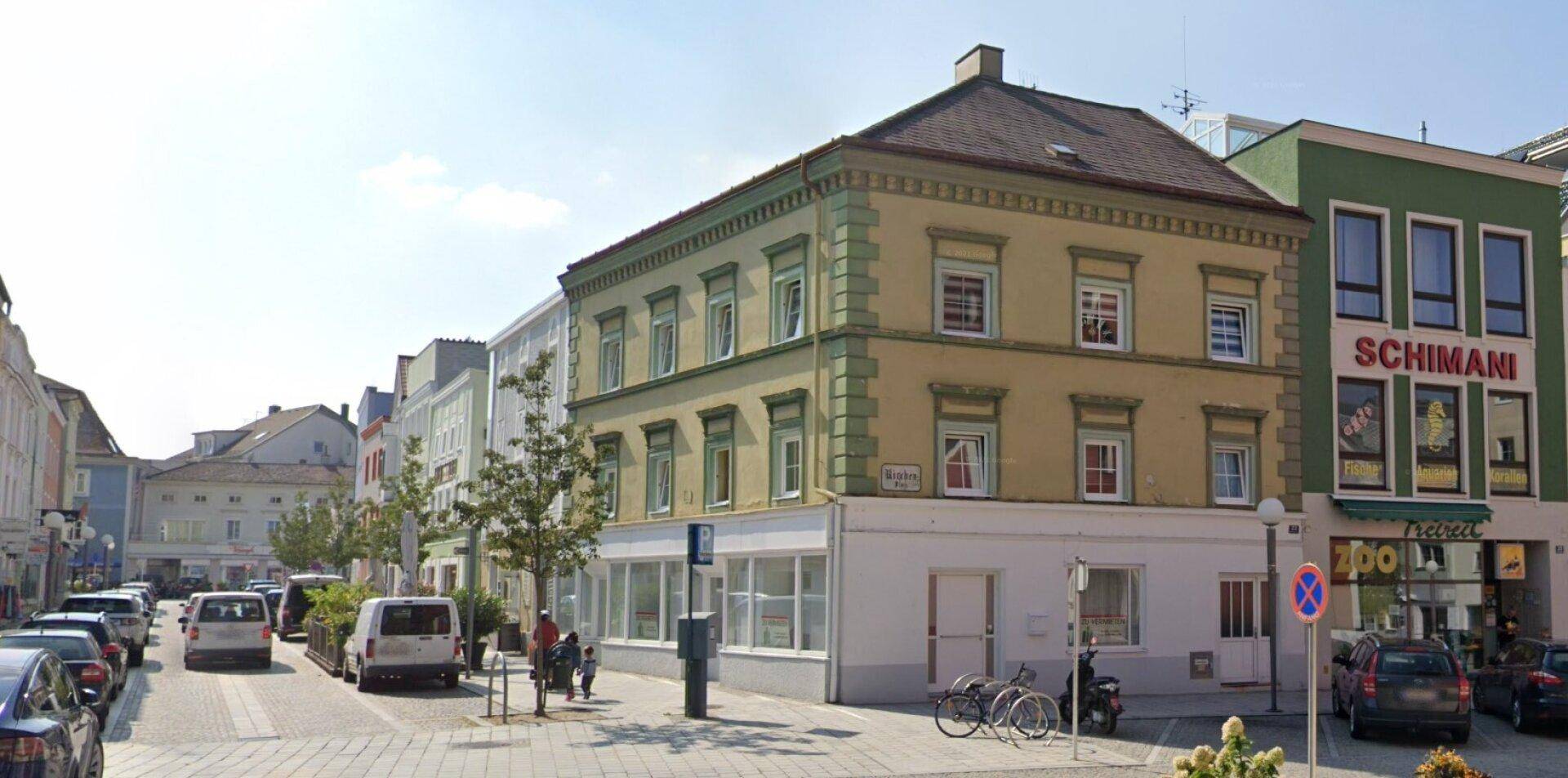 Historisches Eckgebäude an einer belebten Straße mit parkenden Autos und Geschäften im Erdgeschoss.