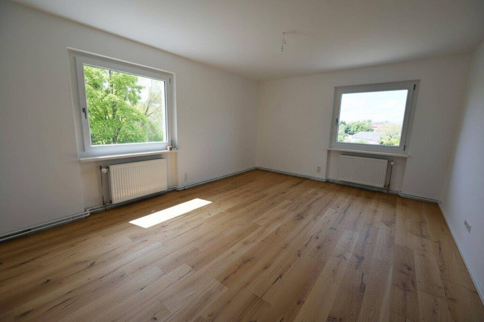 Helles, unmöbliertes Zimmer mit großen Fenstern, Holzboden und viel Tageslicht.