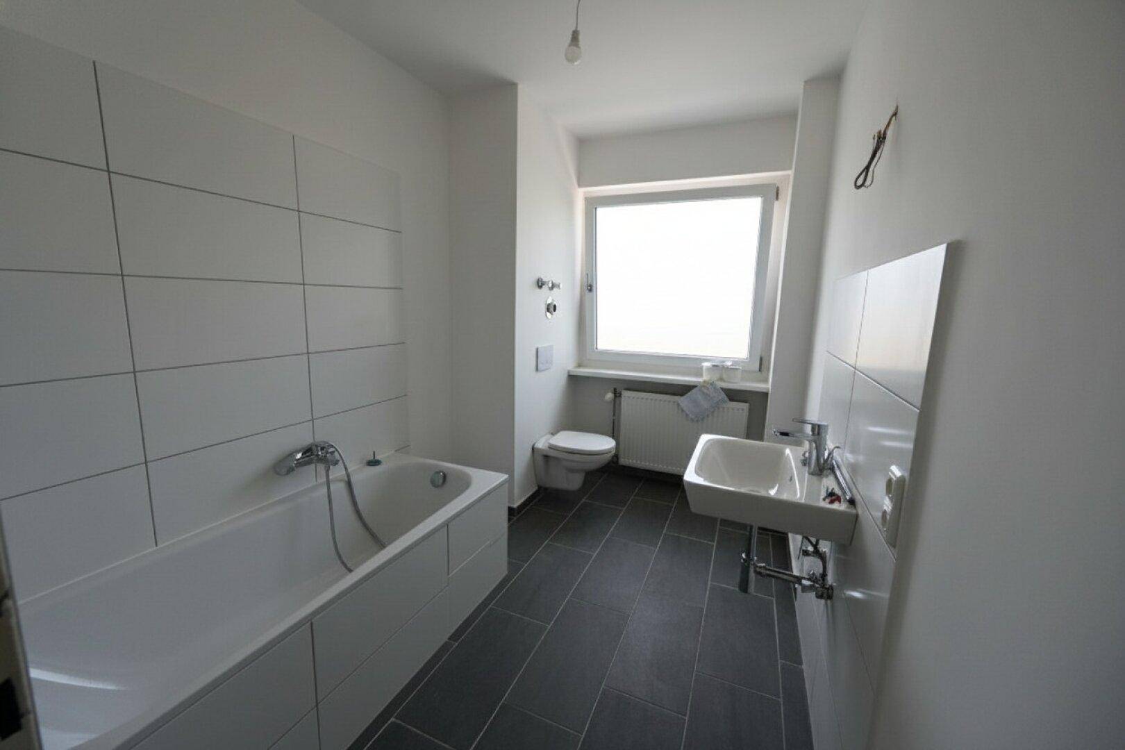 Helles Badezimmer mit Badewanne, WC, Waschbecken und Fenster, ausgestattet mit weißen Fliesen.