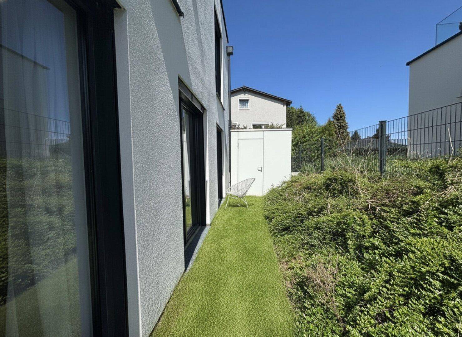 Seitenansicht des Gebäudes mit schmalem Rasenstreifen und grünen Büschen entlang der Fassade.