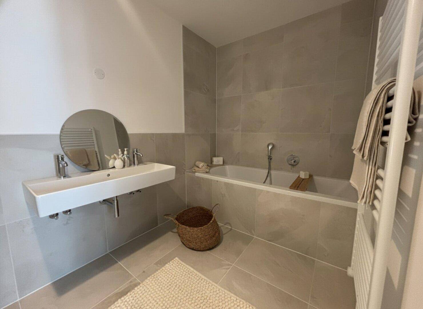 Modernes Badezimmer mit grauen Fliesen, Badewanne und einem stilvollen Doppelwaschbecken.