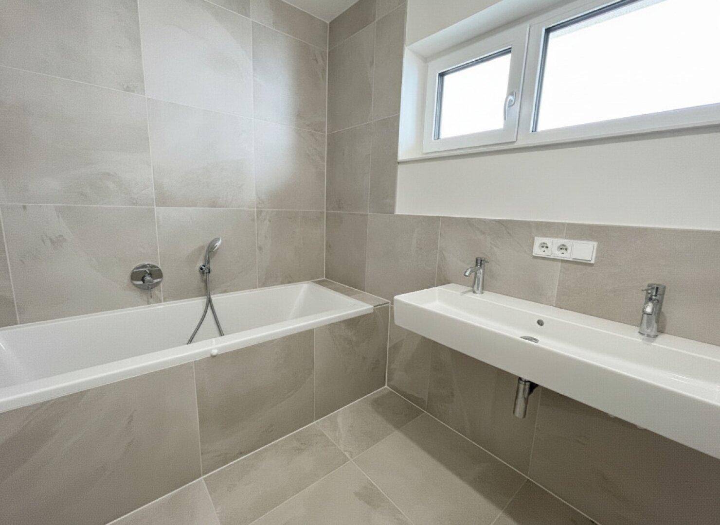 Modernes Badezimmer mit hellgrauen Fliesen, Badewanne und Doppelwaschbecken unter einem Fenster.