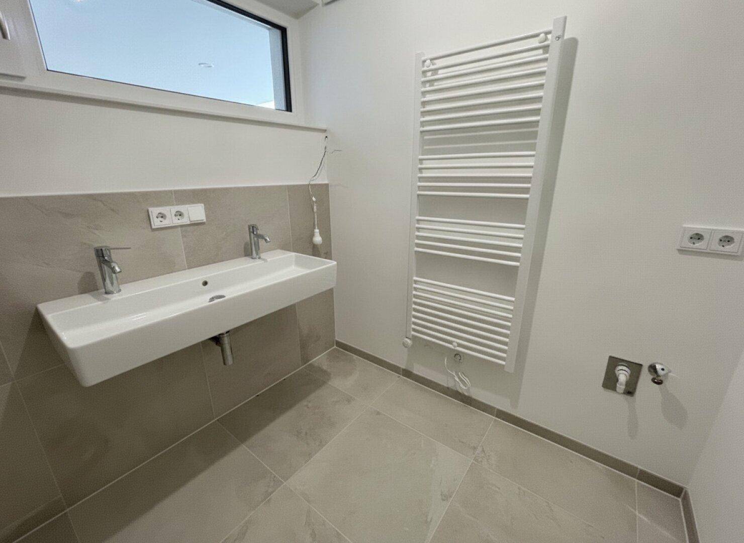 Modernes Badezimmer mit Doppelwaschbecken, beheiztem Handtuchhalter und Fenster, in hellen Grautönen gehalten.