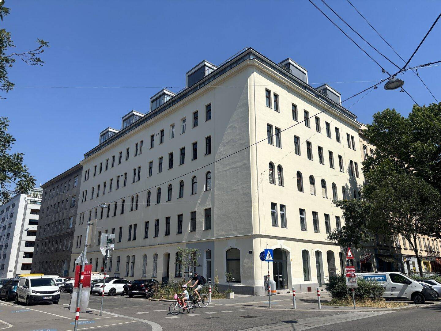 Großes Eckgebäude mit vielen Fenstern an einer belebten Straße unter blauem Himmel.