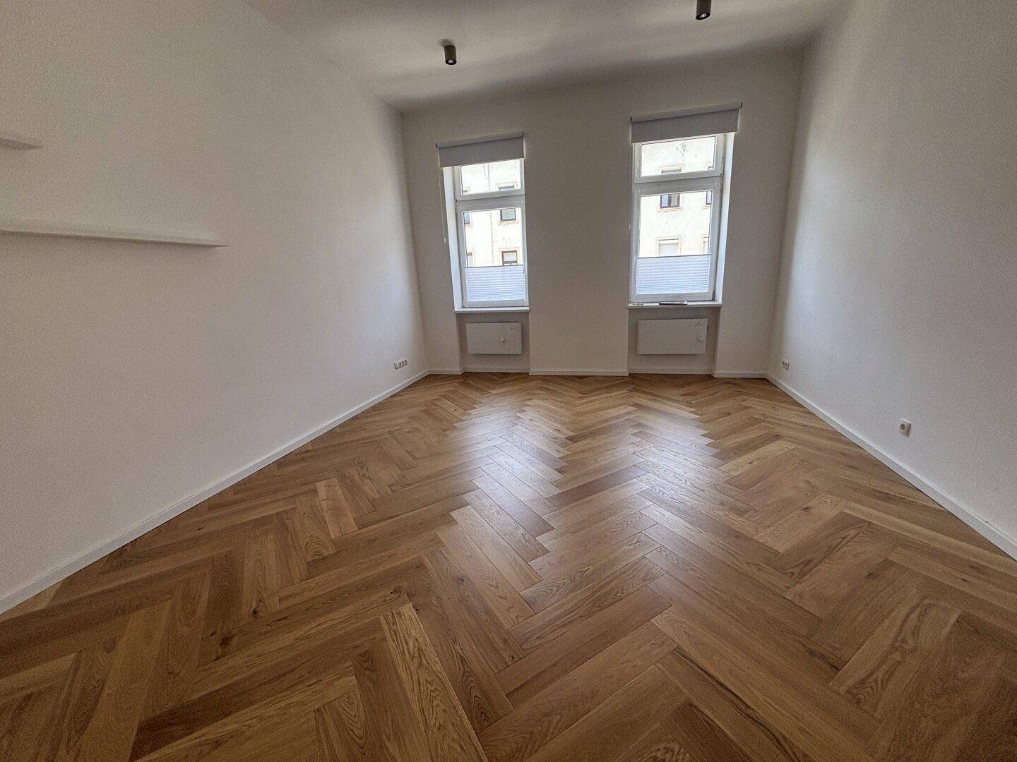 Heller, leerer Raum mit elegantem Fischgrätparkett und zwei Fenstern, bereit zur individuellen Gestaltung.