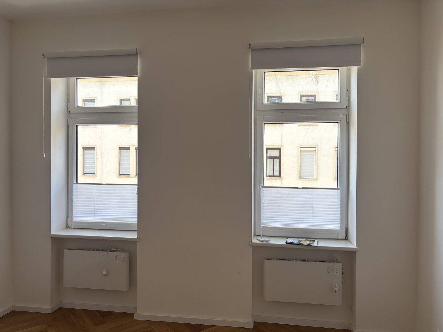 Zwei Fenster mit modernen Rollos und Heizkörpern darunter, die einen Blick auf die umliegenden Gebäude bieten.