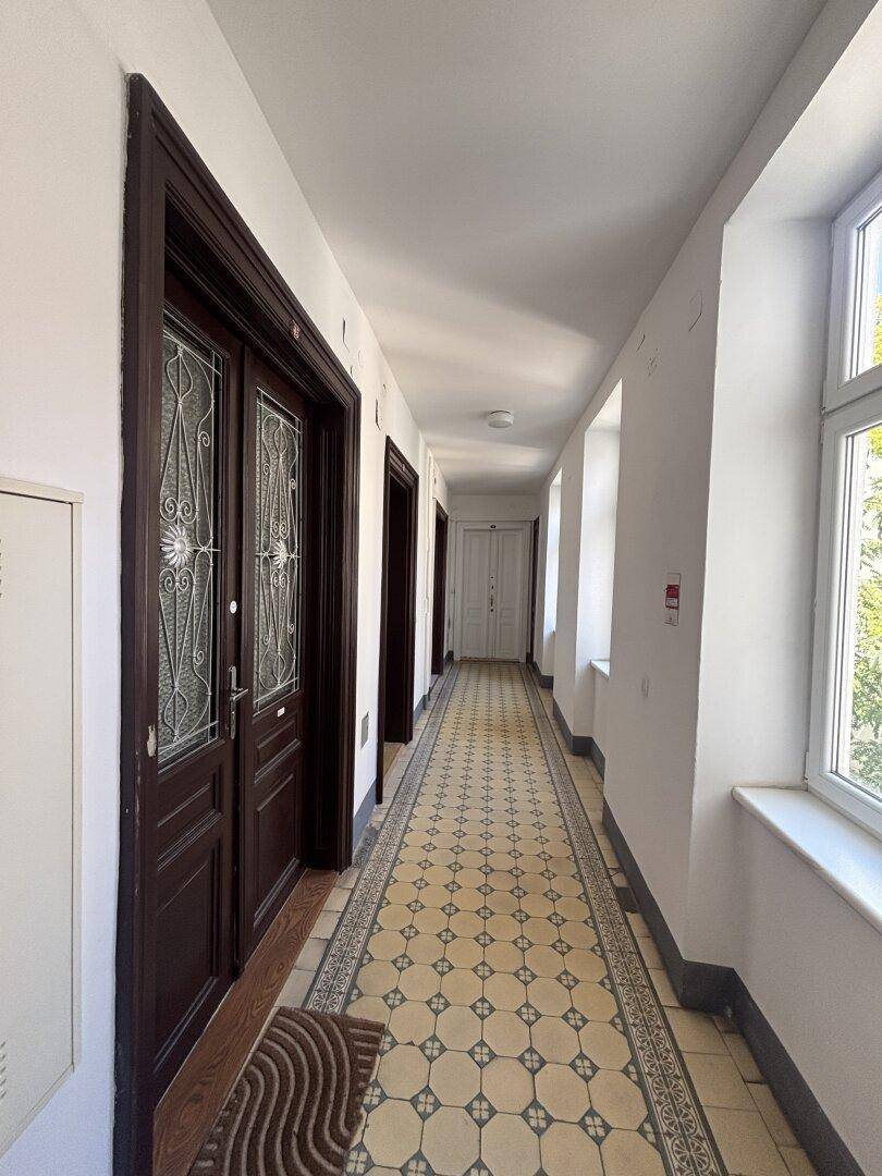 Langer Gemeinschaftsflur mit gemusterten Fliesen, dunklen Holztüren und Fenstern, die Tageslicht hereinlassen.