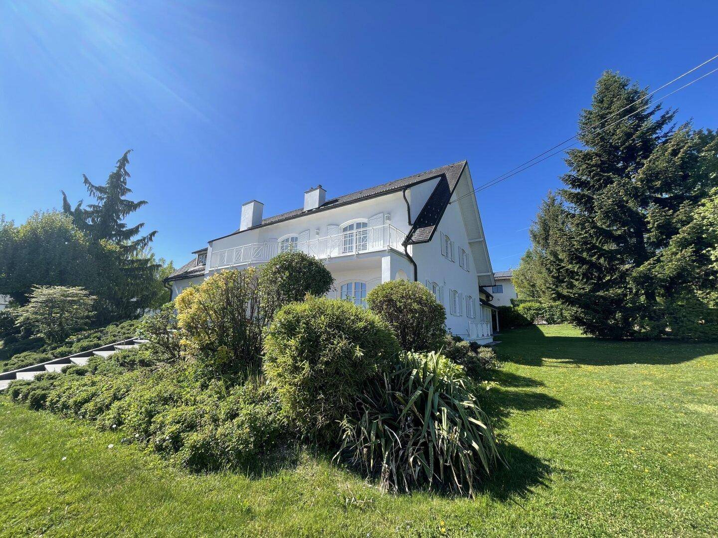 Seitenansicht des weißen Hauses mit üppiger Bepflanzung und großem Gartenbereich unter strahlendem Himmel.