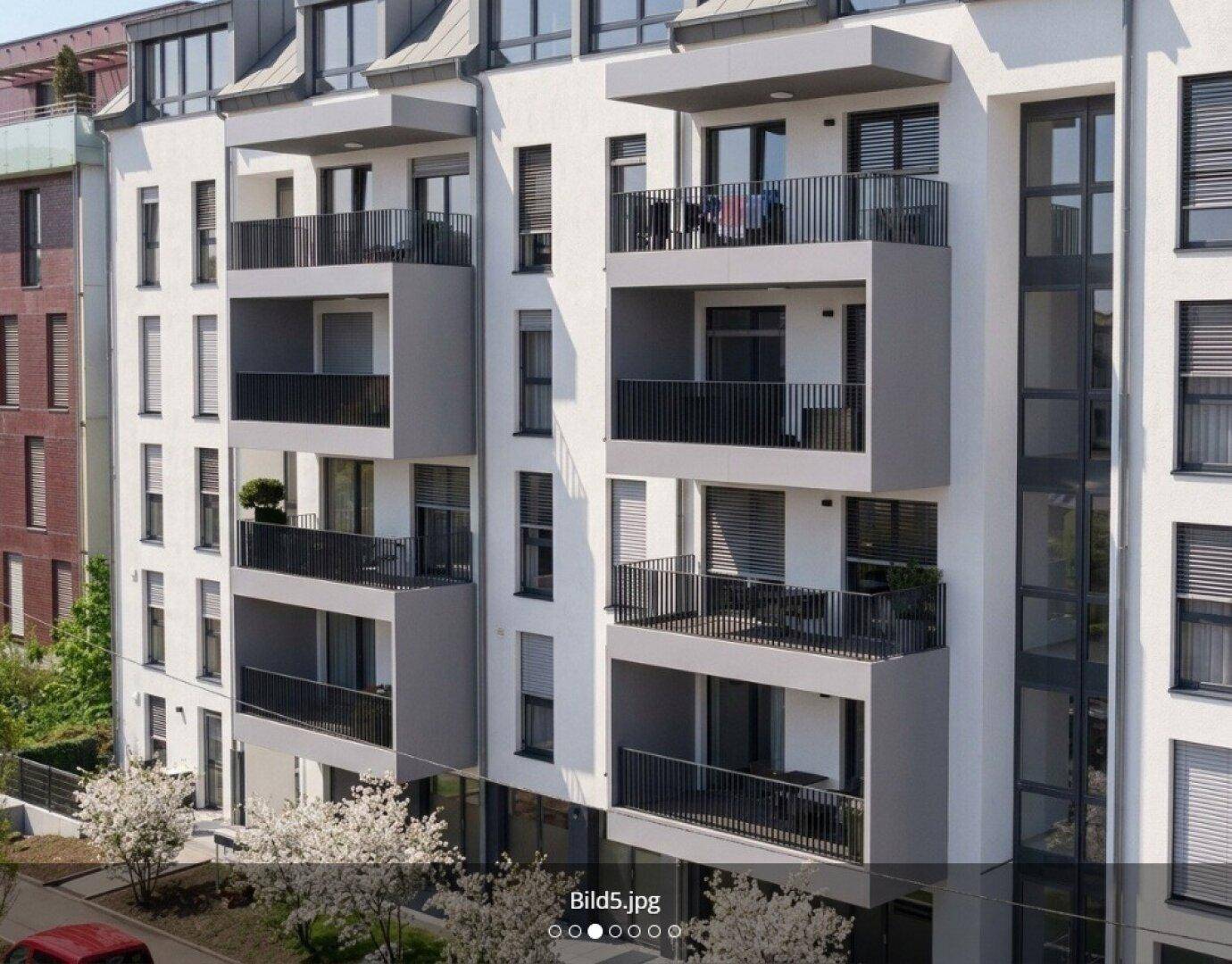 Moderne Wohnhausfassade mit zahlreichen Balkonen und großen Fenstern, umgeben von blühenden Bäumen.