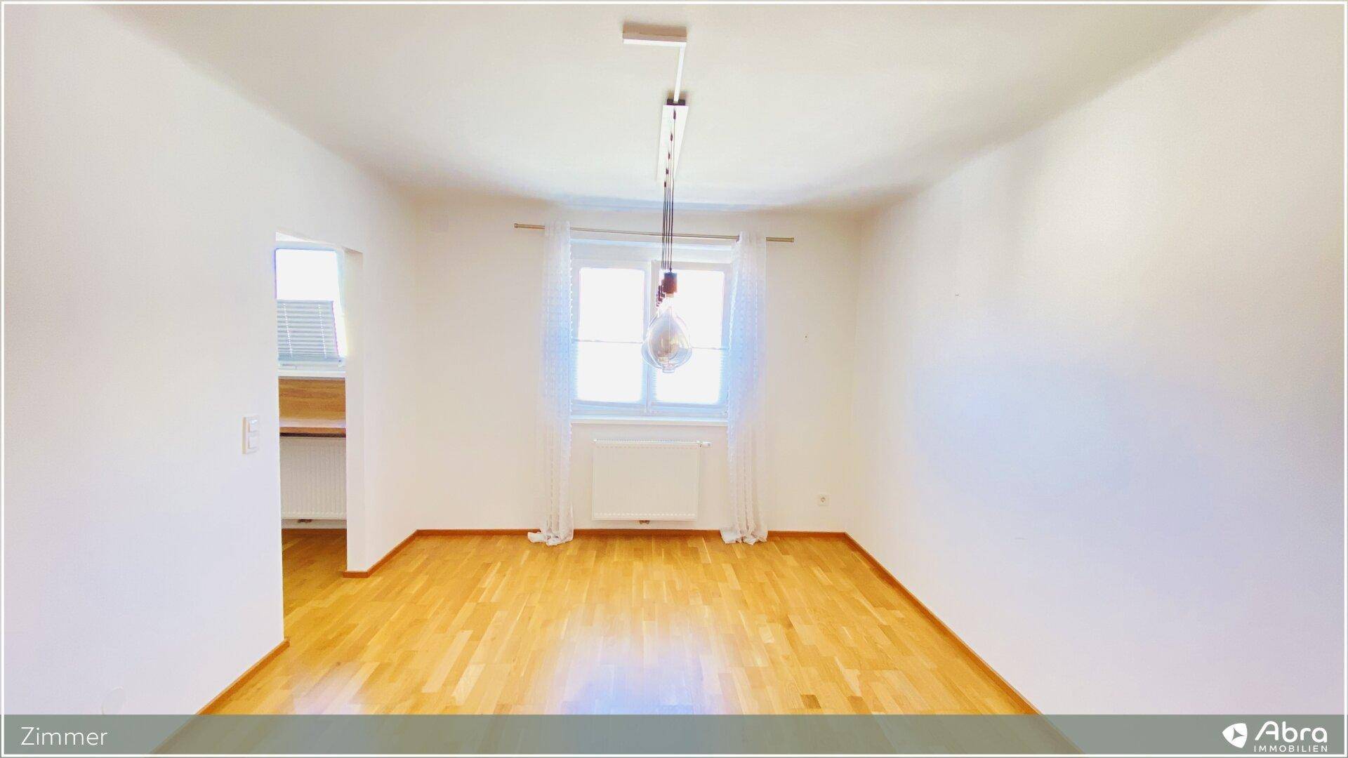 Leeres Zimmer mit Holzboden und einem Fenster, das viel Tageslicht hereinlässt.