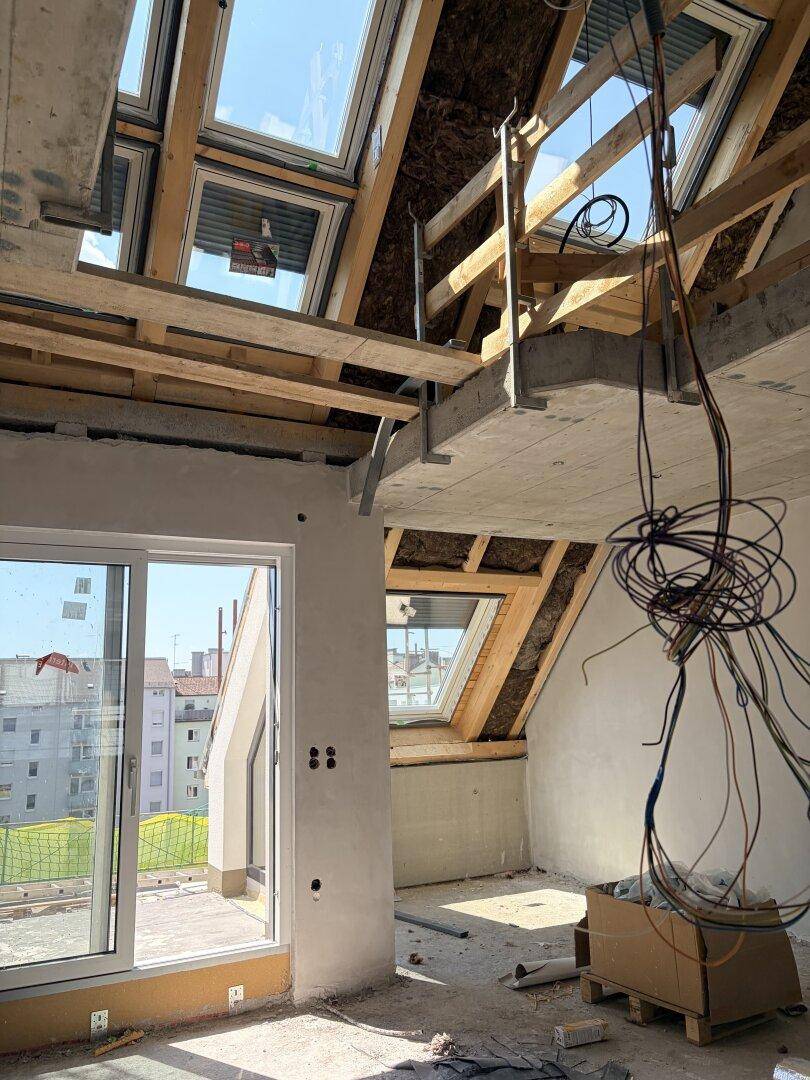 Unfertiger Innenraum im Dachgeschoss mit großen Fenstern, sichtbaren Decken und Blick ins Freie.