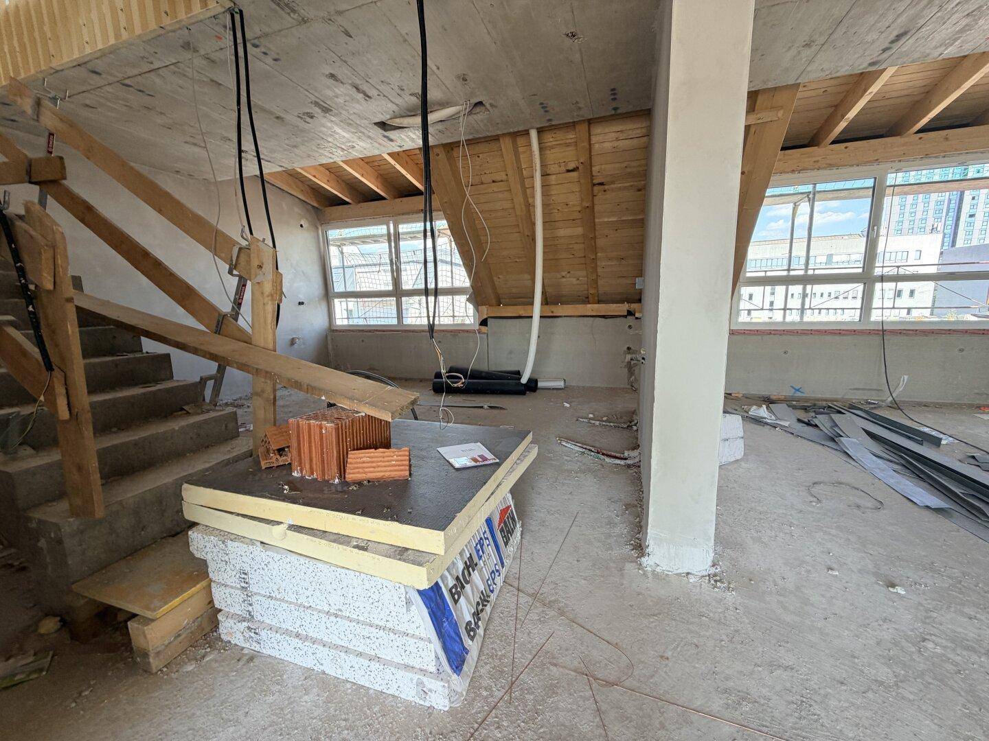 Baustelle eines Innenraums mit Treppe, sichtbaren Holzdeckenbalken und Fenstern.