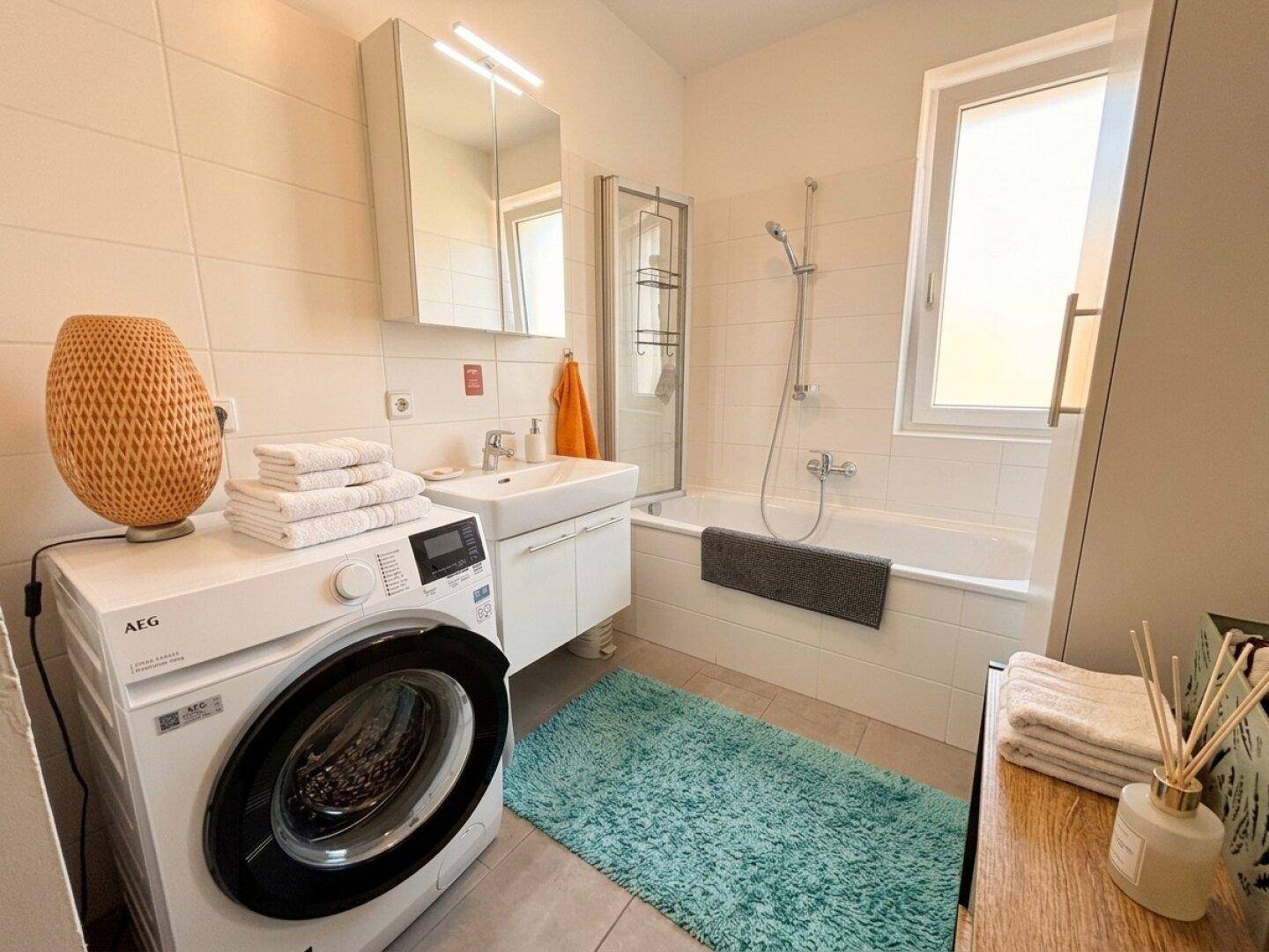 Helles Badezimmer mit Badewanne, Dusche, Waschbecken, Waschmaschine und Fenster.