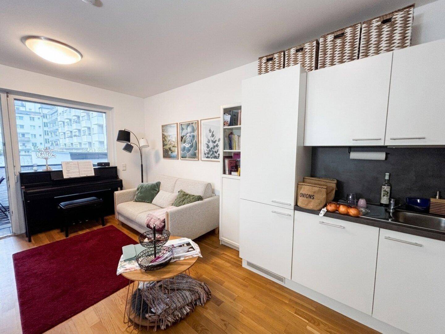 Wohnbereich mit integrierter Küchenzeile, Sofa, Klavier und Zugang zum Balkon.