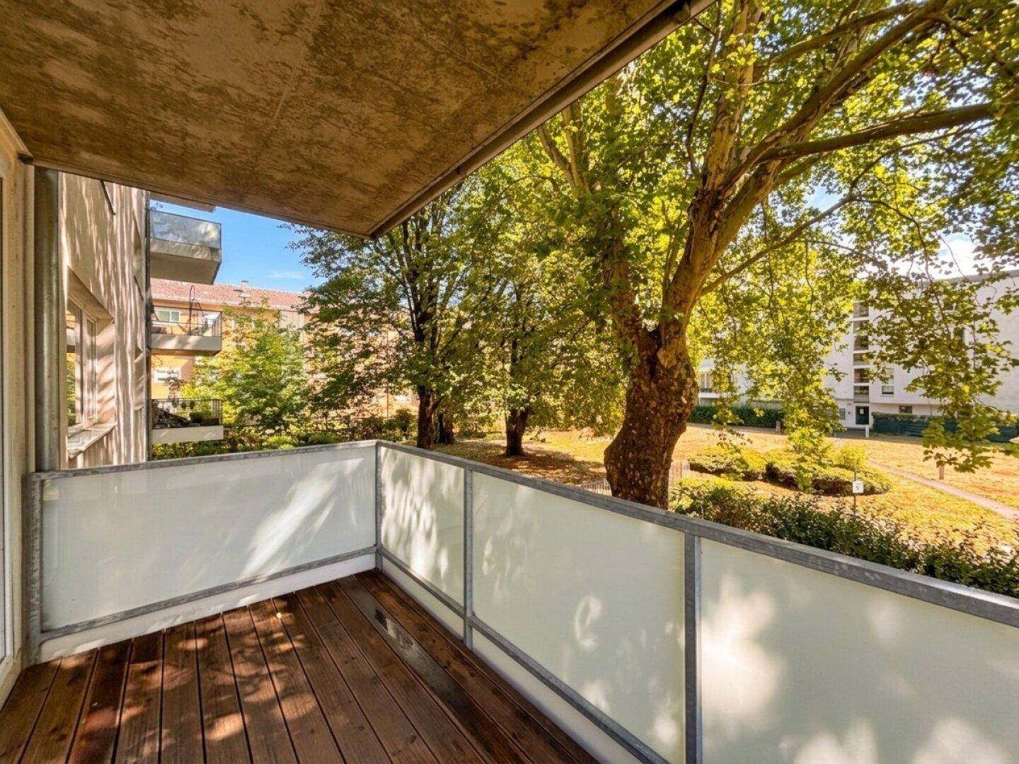 Geräumiger Balkon mit Holzboden und Blick auf den begrünten Innenhof mit hohen Bäumen.