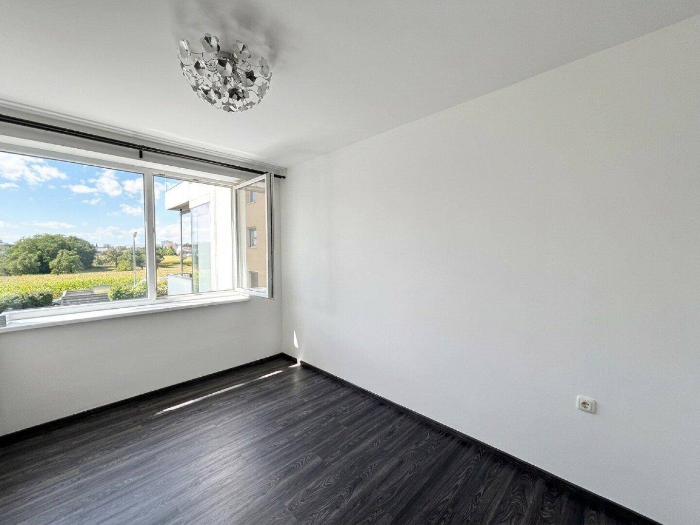 Helles Zimmer mit dunklem Holzboden und weitem Blick ins Grüne durch ein großes Fenster.