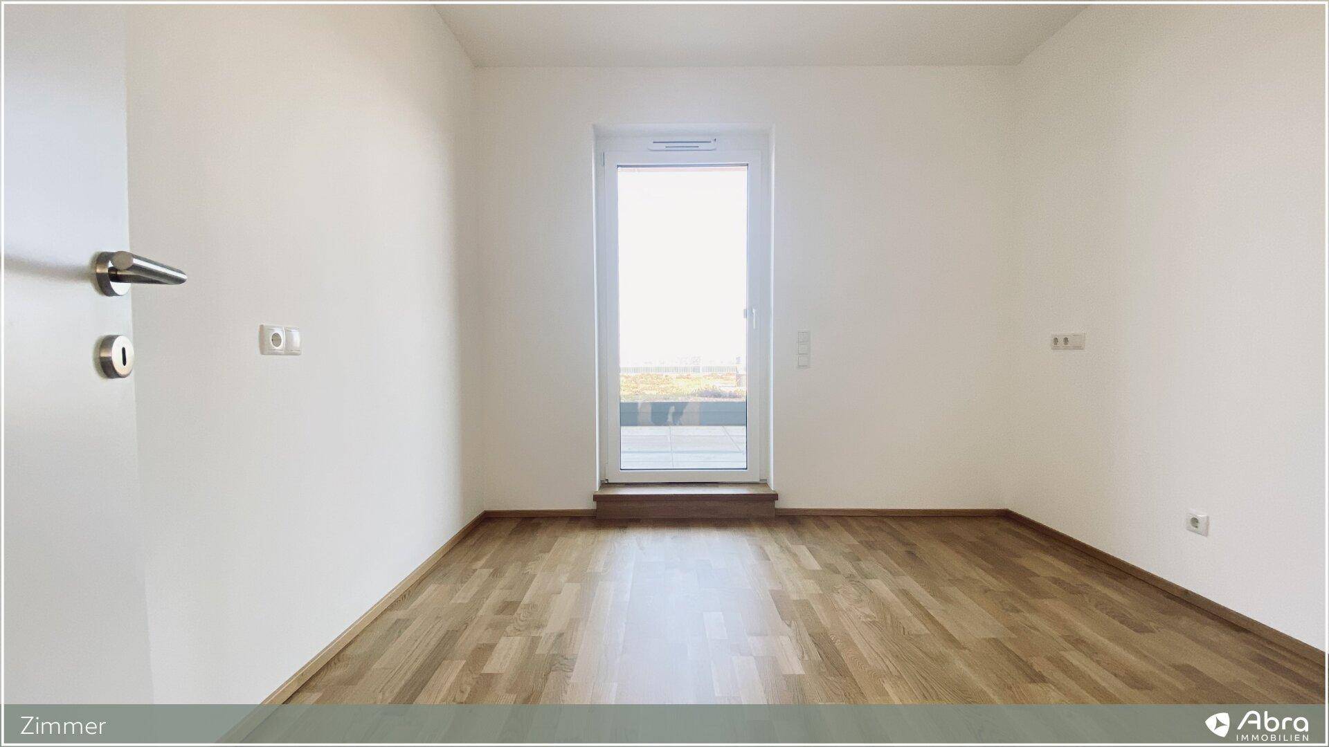 Leerer Raum mit Parkettboden und großem Fenster, das viel Tageslicht hereinlässt.