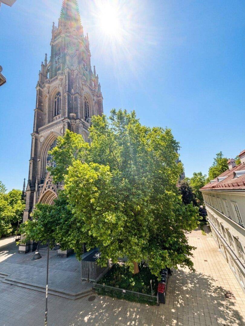 Beeindruckende gotische Kirche mit hohem Turm, umgeben von grünen Bäumen unter strahlend blauem Himmel.